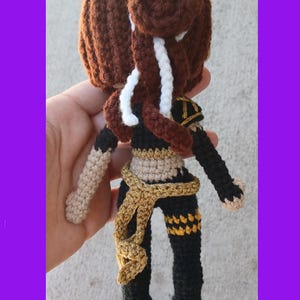 May include: Crocheted doll with brown hair styled in a bun and braids, wearing a black and gold outfit. The doll has tan arms and a gold sash. The doll is handmade and has a unique design.