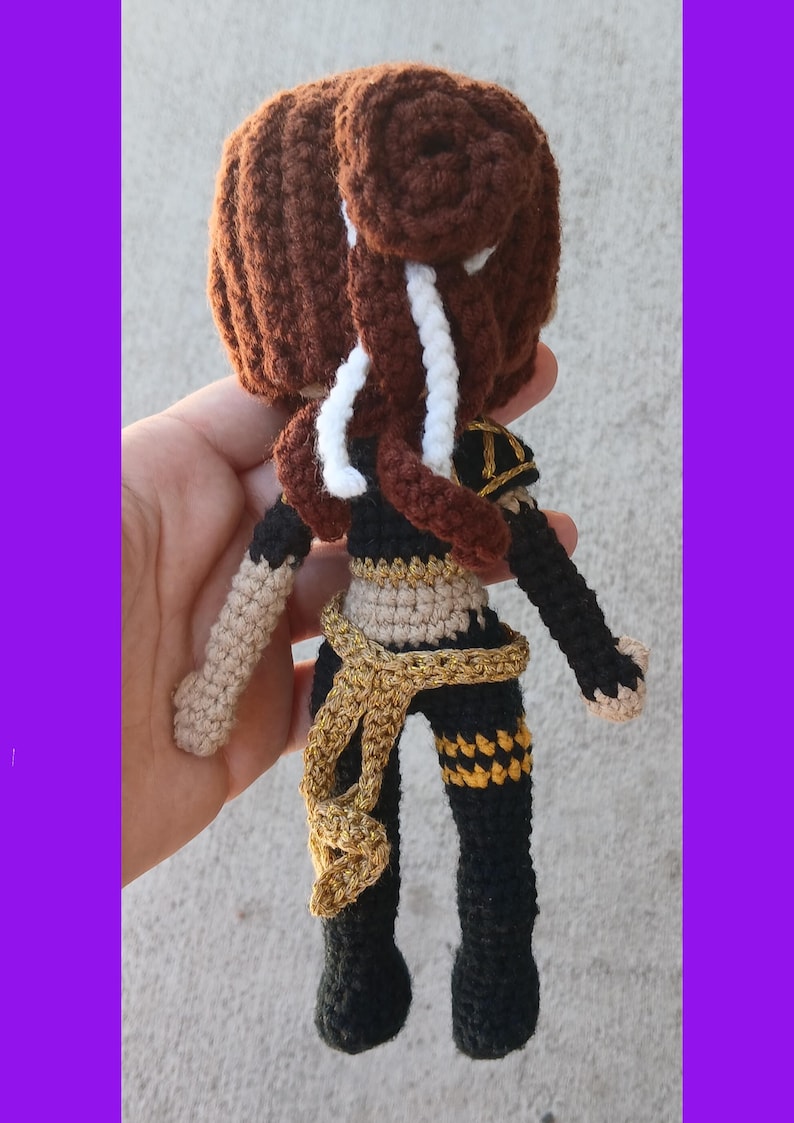 May include: Crocheted doll with brown hair styled in a bun and braids, wearing a black and gold outfit. The doll has tan arms and a gold sash. The doll is handmade and has a unique design.