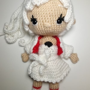 May include: A crocheted doll with white hair, a white dress, and red accents. The doll has black eyes and a sweet expression.