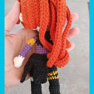 May include: A handmade crochet doll with orange hair and a black outfit. The doll has a purple sleeve with a yellow star design. The doll is held in a hand.
