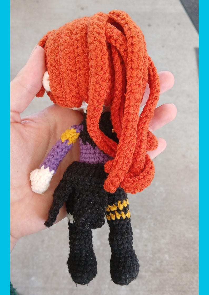 May include: A handmade crochet doll with orange hair and a black outfit. The doll has a purple sleeve with a yellow star design. The doll is held in a hand.