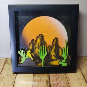 May include: A framed 3D art piece depicting a desert landscape with a large orange moon, silhouetted mountains, and green cacti. The artwork is set against a black background.