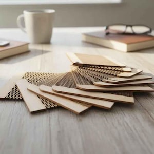 Puede incluir: Una colecci&oacute;n de muestras de madera cortadas con l&aacute;ser con varios patrones, abanicadas sobre una superficie de madera clara. Tambi&eacute;n se ven una taza blanca, dos cuadernos encuadernados en cuero y un par de gafas, lo que sugiere un espacio de trabajo o estudio de dise&ntilde;o.