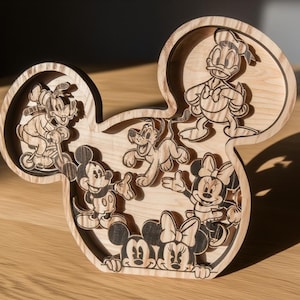 Può includere: Elemento decorativo in legno a forma di testa di Topolino, con immagini incise di personaggi Disney. I personaggi includono Topolino, Minnie, Paperino, Pippo e Pluto. Il pezzo è realizzato in legno chiaro e poggia su una superficie di legno.