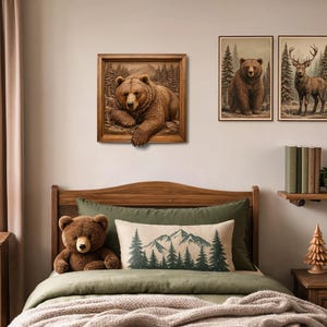 May include: A square framed wall art piece featuring a three-dimensional brown bear resting in a forest setting, with the bear's paw extending beyond the frame. The artwork is displayed on a wall above a bed with green bedding and decorative pillows.