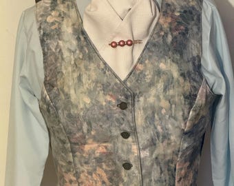 Puede incluir: Un chaleco con un patrón de acuarela en tonos grises, azules y rosas. Tiene cuello en V y botones. Debajo se lleva un ascot blanco, asegurado con un broche rosa. Se ve una camisa azul claro debajo.
