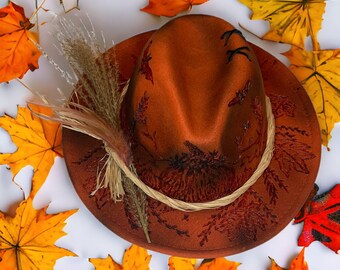 May include: A brown felt hat with a wide brim and a decorative woven band. The hat has a detailed design of leaves and branches in a darker brown color. The hat is surrounded by autumn leaves.