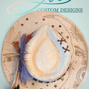 May include: A cream-colored custom design hat with a decorative brim featuring a celestial design. The hat has a light-colored crown with brown stitching and is adorned with blue and brown feathers. The brim is decorated with stars, moons, and a face.