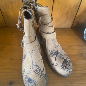 May include: A pair of brown suede ankle boots with black dragonfly and flower designs. The boots have laces and a zipper on the side.