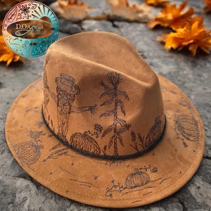 May include: A brown felt fedora hat with a black band and a hand-drawn design of a scarecrow, corn stalks, and pumpkins.