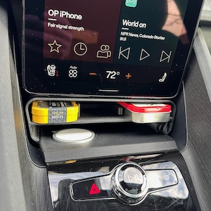Könnte beinhalten: Ein schwarzes Armaturenbrett mit einem Bildschirm, der "OP iPhone" und "World on" mit einer Temperaturanzeige von 72 Grad anzeigt. Unter dem Bildschirm befindet sich ein kleines weißes kabelloses Ladegerät und zwei kleine rechteckige Behälter, einer gelb und einer rot, im Ablagefach.