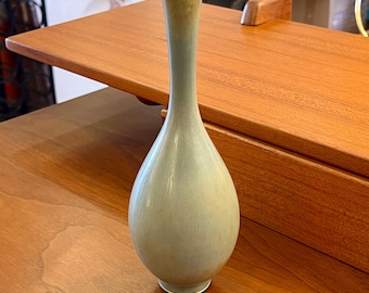 Puede incluir: Un jarrón de cerámica azul claro con fondo redondeado y cuello estrecho. El jarrón está sobre una superficie de madera.