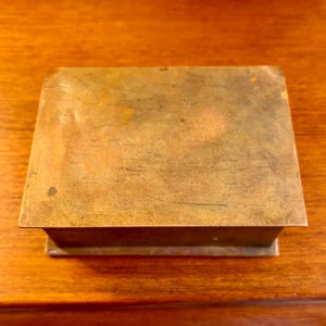 May include: A rectangular brass box with a worn, aged finish. The box has a simple design with a flat top and a slightly raised edge.