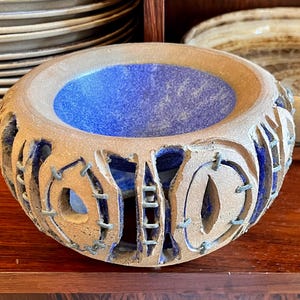 May include: A handcrafted ceramic bowl with a blue interior and a textured, beige exterior. The bowl features cut-out designs and is displayed on a wooden surface. Other ceramic items are visible in the background.