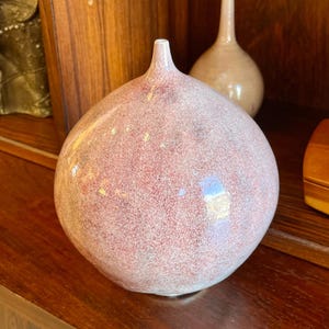 Puede incluir: Un jarrón redondo de cerámica rosa moteado con un cuello estrecho. El jarrón tiene forma bulbosa y superficie texturizada. En el fondo se ve un jarrón más pequeño, beige. Los jarrones se exhiben sobre una superficie de madera oscura.