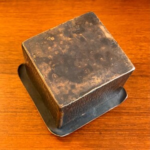 May include: A square, dark brown metal box with a textured surface and a slightly raised rim. The box has a slightly worn appearance.