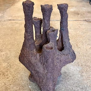 May include: A textured, dark purple ceramic sculpture with multiple vertical, cylindrical forms. The sculpture has a rough, organic appearance, with a base that appears to be a solid mass. The piece is likely a decorative item.