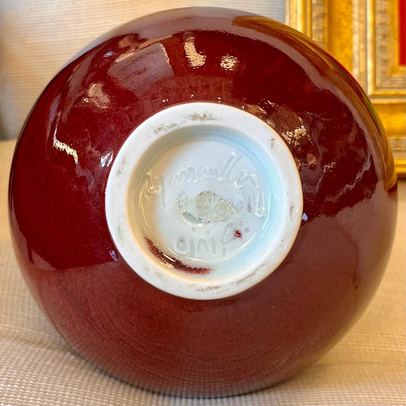 May include: A round, burgundy-colored ceramic vase with a glossy finish. The base is white with script text and a signature. The vase has a smooth, curved shape, and the color is consistent throughout.