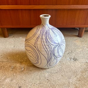 May include: A white ceramic vase with a blue abstract design. The vase has a round body and a narrow neck.