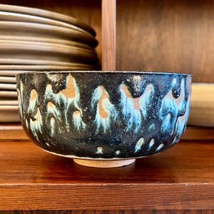 May include: A ceramic bowl with a dark blue and black glaze, featuring a pattern of gold and turquoise accents. The bowl is round with a slightly flared rim and sits on a small, unglazed base. The bowl is displayed on a wooden surface.