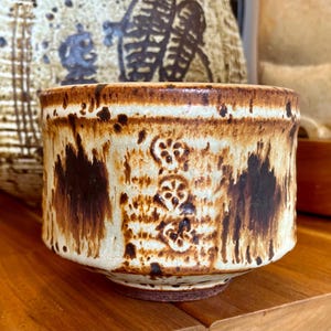 Könnte beinhalten: Eine Keramikschale mit cremefarbenem Grund und braunen Akzenten. Die Schale hat eine strukturierte Oberfläche mit dunkelbraunen Tropfenmustern und drei floralen Designs. Der Rand und der Boden sind dunkler braun. Die Schale steht auf einer Holzoberfläche.