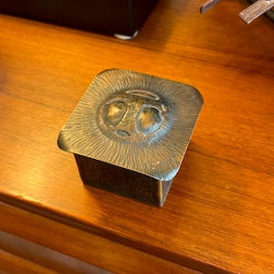 May include: A square, bronze trinket box with a textured surface and a raised, embossed design of a face with two eyes.