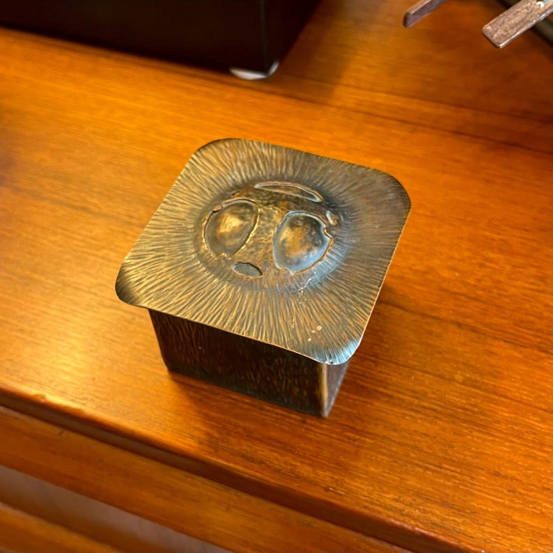 May include: A square, bronze trinket box with a textured surface and a raised, embossed design of a face with two eyes.
