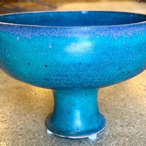 May include: A teal blue ceramic bowl with a wide, rounded base and a flared lip. The bowl has a speckled finish and a slightly iridescent glaze.