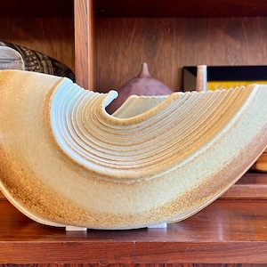 May include: A ceramic decorative bowl with a unique, curved design. The bowl features a light tan color with darker brown accents along the edges. The interior has a series of curved ridges, adding texture and visual interest. The bowl is a decorative item.