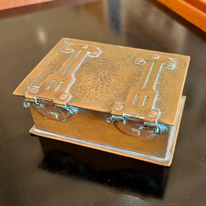 May include: A small, rectangular copper box with a hammered finish and intricate geometric designs on the lid. The box has a hinged lid and is decorated with a green patina.
