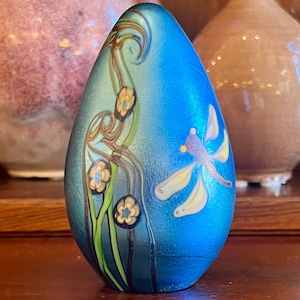 May include: A blue glass egg-shaped decorative object with a dragonfly and floral design. The egg features a textured surface and is adorned with a dragonfly and floral pattern in shades of yellow, green, and brown. The background is a gradient of blue.