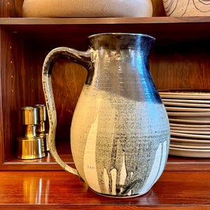 May include: A handcrafted ceramic pitcher with a large handle. The pitcher features a two-tone design, with a dark brown top and a cream-colored base with black dripped accents. The pitcher is displayed on a wooden shelf.