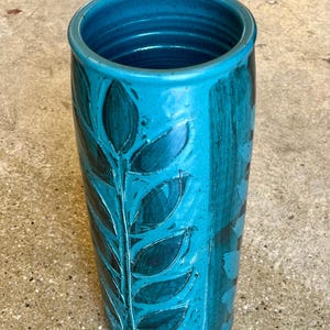 May include: A tall, cylindrical turquoise vase with a textured surface. The vase features a hand-painted design of dark teal leaves and stems. The interior rim has a darker teal color. A decorative piece for the home.