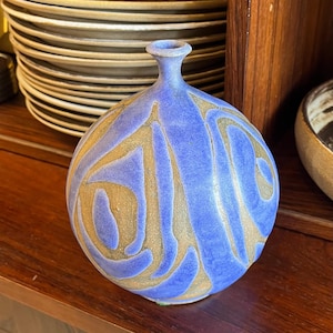 May include: A blue ceramic vase with a bulbous body and a narrow neck. The vase features a swirling pattern in shades of gold. The vase is displayed on a wooden surface, with a stack of plates in the background.