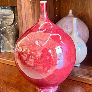 May include: A glossy, red decorative vase with a bulbous body and a slender neck. The vase has a reflective surface, with white accents and a pink base. Other vases are visible in the background, sitting on a wooden surface.