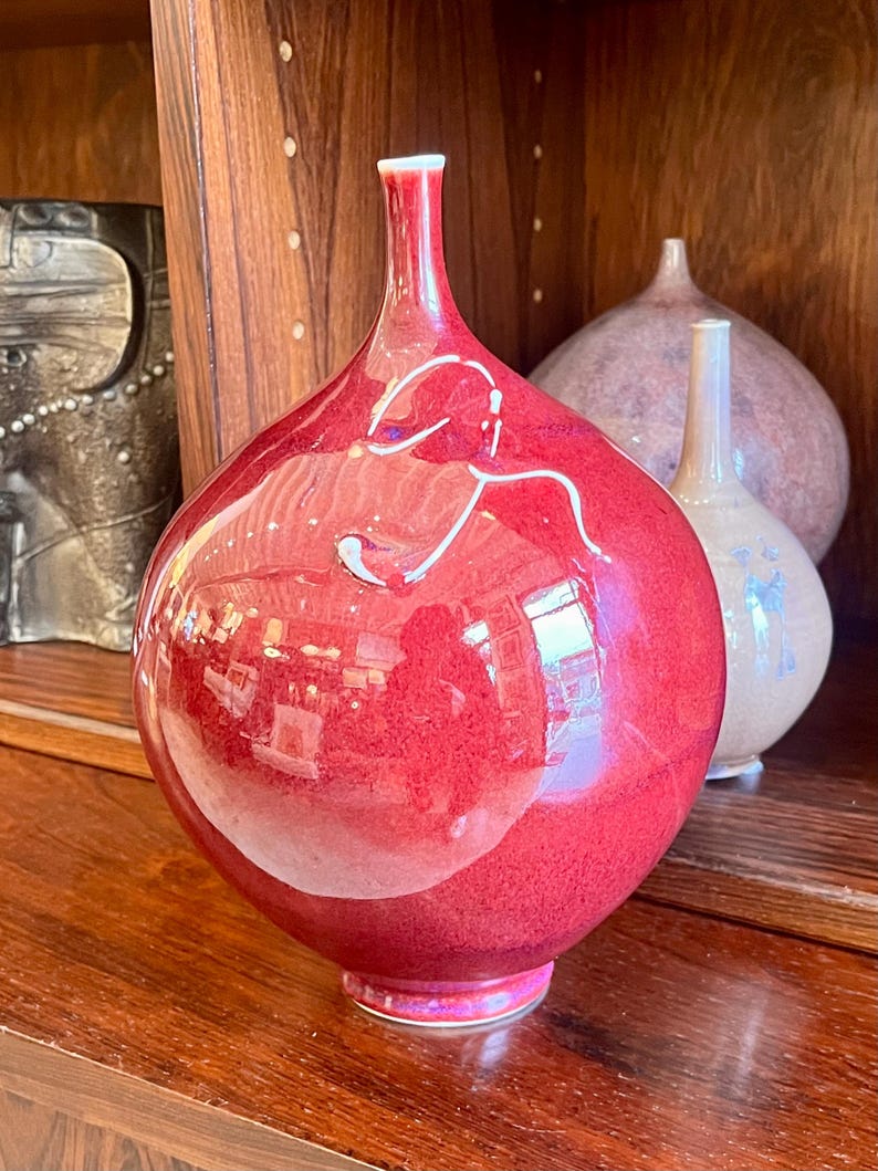 May include: A glossy, red decorative vase with a bulbous body and a slender neck. The vase has a reflective surface, with white accents and a pink base. Other vases are visible in the background, sitting on a wooden surface.