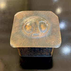 May include: A square copper box with a hammered finish. The lid features a raised design of a stylized face with two large eyes.