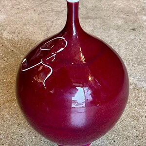 May include: A glossy, burgundy-colored ceramic vase with a bulbous body and a slender neck. The vase features a white, abstract design on its surface. The base has a pink accent, and the rim is white.