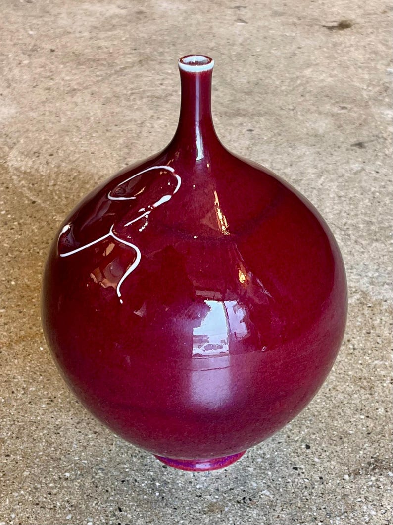 May include: A glossy, burgundy-colored ceramic vase with a bulbous body and a slender neck. The vase features a white, abstract design on its surface. The base has a pink accent, and the rim is white.