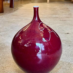 May include: A glossy, maroon-colored vase with a bulbous body and a slender neck. The vase features a white rim and a decorative white line design. The base is a darker shade of maroon. The vase is a decorative home decor item.