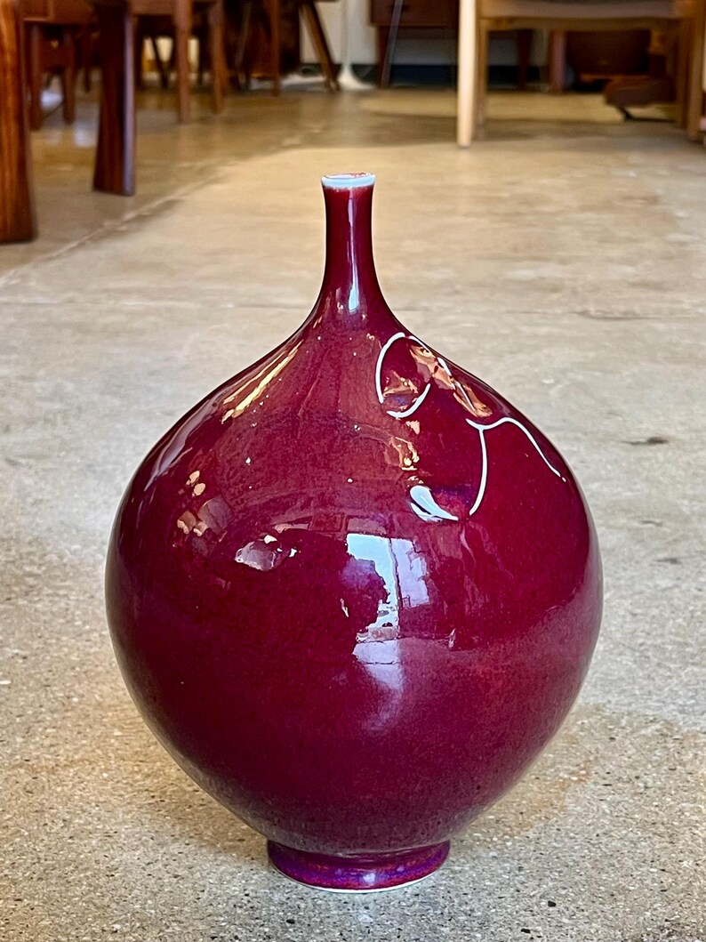 May include: A glossy, maroon-colored vase with a bulbous body and a slender neck. The vase features a white rim and a decorative white line design. The base is a darker shade of maroon. The vase is a decorative home decor item.