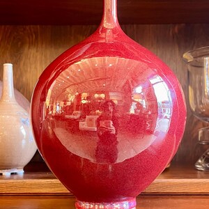 May include: A large, glossy red vase with a bulbous body and a slender neck. The vase has a textured surface and a white rim. It is displayed on a wooden shelf, reflecting the surrounding environment.
