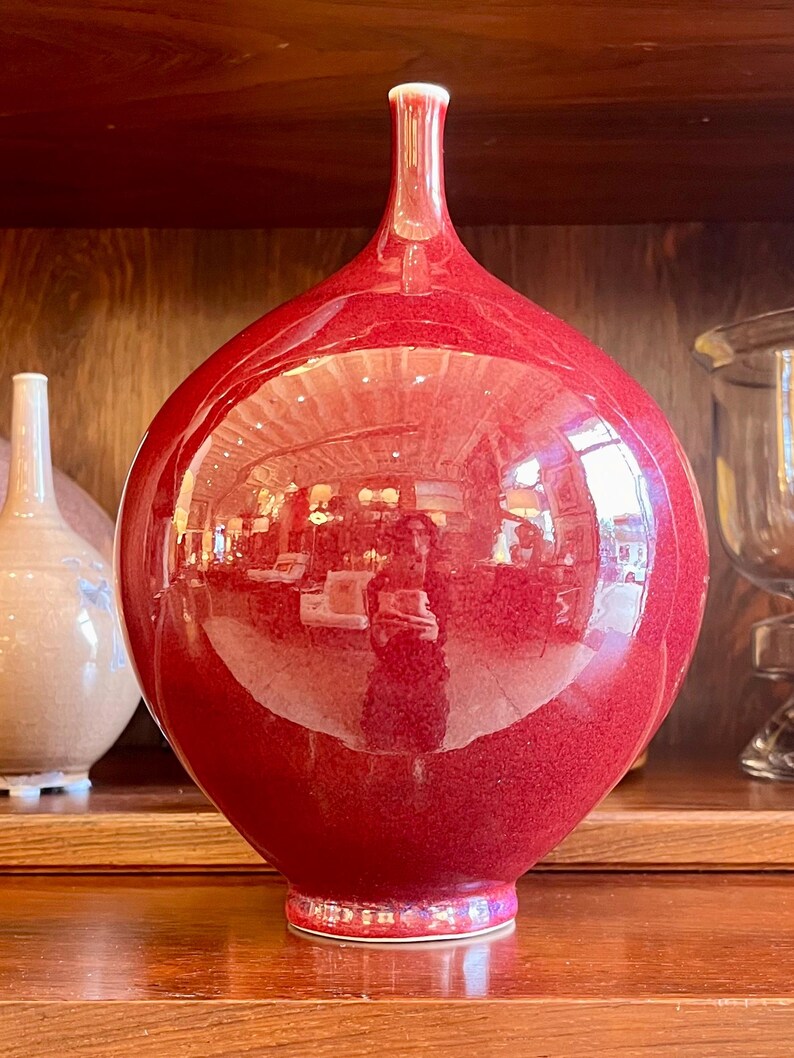 May include: A large, glossy red vase with a bulbous body and a slender neck. The vase has a textured surface and a white rim. It is displayed on a wooden shelf, reflecting the surrounding environment.