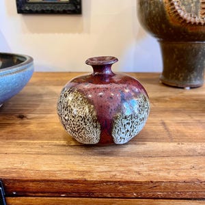 Puede incluir: Un pequeño jarrón redondo de cerámica con un esmalte moteado en tonos marrones, blancos y rojos. El jarrón tiene un cuello estrecho y un labio ligeramente acampanado.