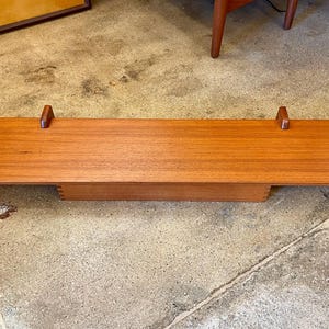 Puede incluir: Una estantería rectangular de madera de estilo moderno de mediados de siglo con un cálido acabado marrón rojizo. Dos pequeños soportes verticales de madera se encuentran en cada extremo. La estantería se encuentra sobre una base rectangular inferior.