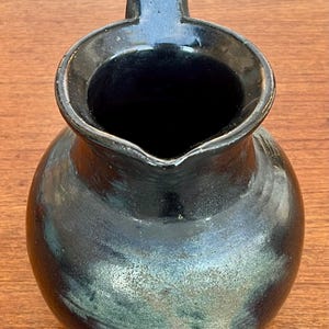 May include: A handcrafted, dark-glazed ceramic pitcher with a rounded body and a sturdy handle. The pitcher has a glossy finish with subtle green and blue hues, adding depth to its dark color. The opening is wide, and the pitcher sits on a wooden surface.