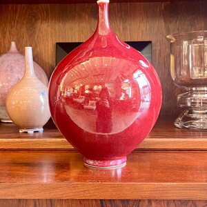May include: A large, glossy red vase with a narrow neck and rounded body. The vase reflects the surrounding environment. Other vases are visible in the background, along with a glass candle holder. The vase is displayed on a wooden shelf.