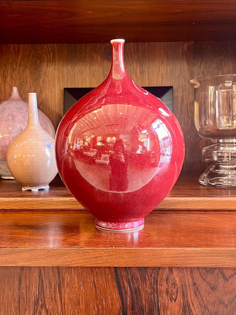 May include: A large, glossy red vase with a narrow neck and rounded body. The vase reflects the surrounding environment. Other vases are visible in the background, along with a glass candle holder. The vase is displayed on a wooden shelf.
