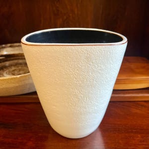 May include: A white textured vase with a black interior and a brown rim. The vase has a unique, slightly tapered shape. The vase is likely ceramic and is displayed on a wooden surface.