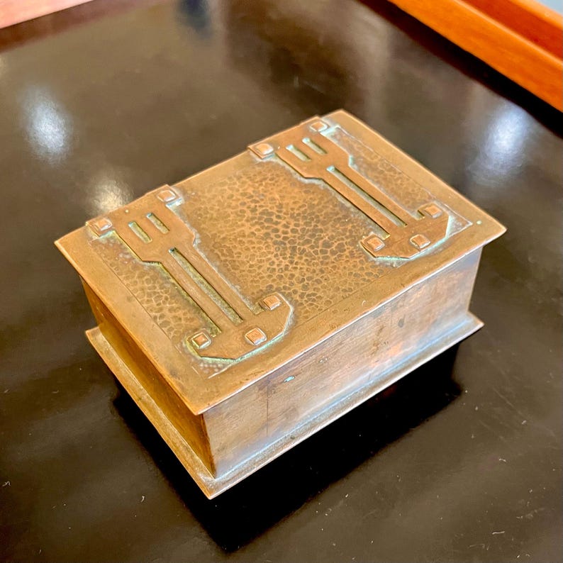 May include: A small, rectangular copper box with a hammered finish and an intricate geometric design. The box has a hinged lid and is decorated with raised squares and lines.